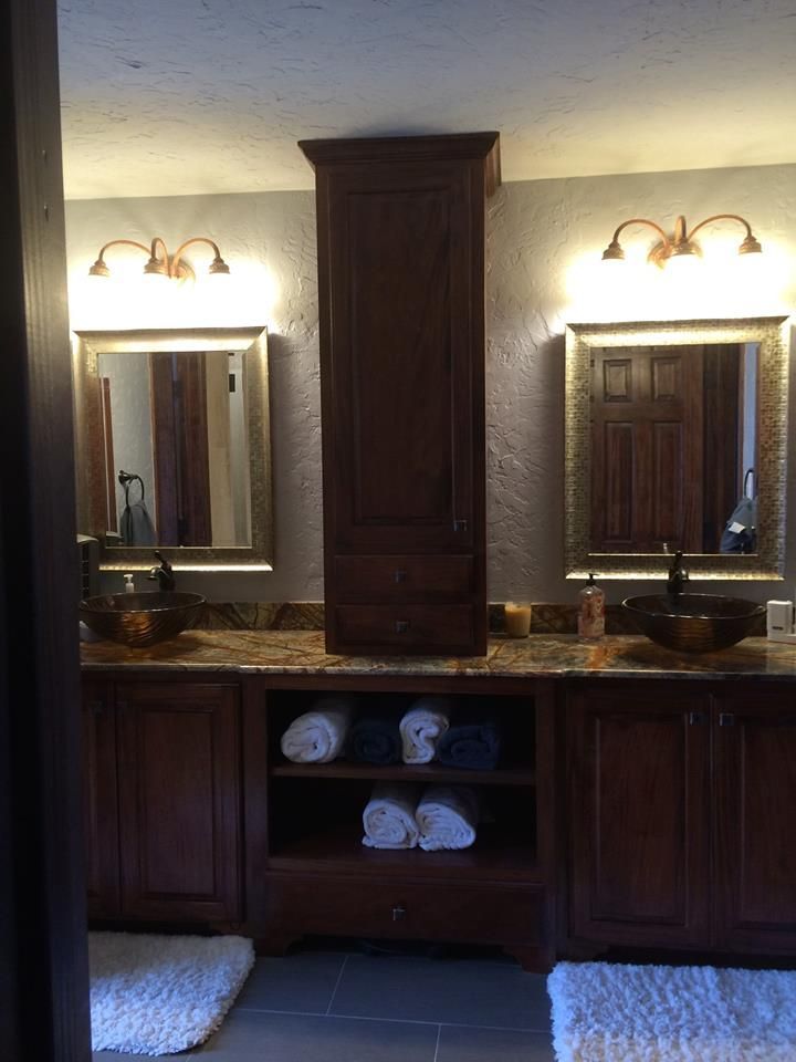 Bathroom with two vanities, each with a mirror, sink, and sconce lights. A tall cabinet stands between them.