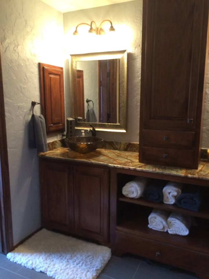 Bathroom with brown cabinetry, granite countertop, glass sink, mirror, and shelves with towels.