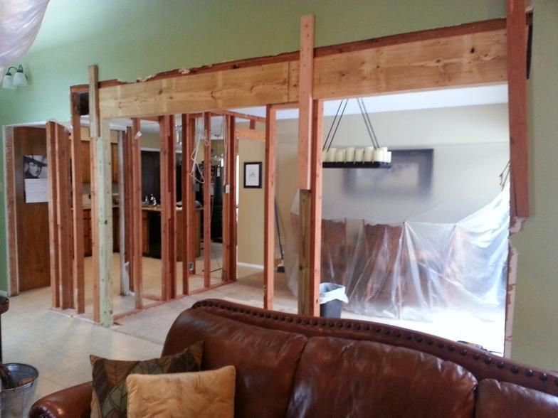 Interior wall demolition, showing wooden framing and opening into a dining room; plastic sheeting covers furniture.