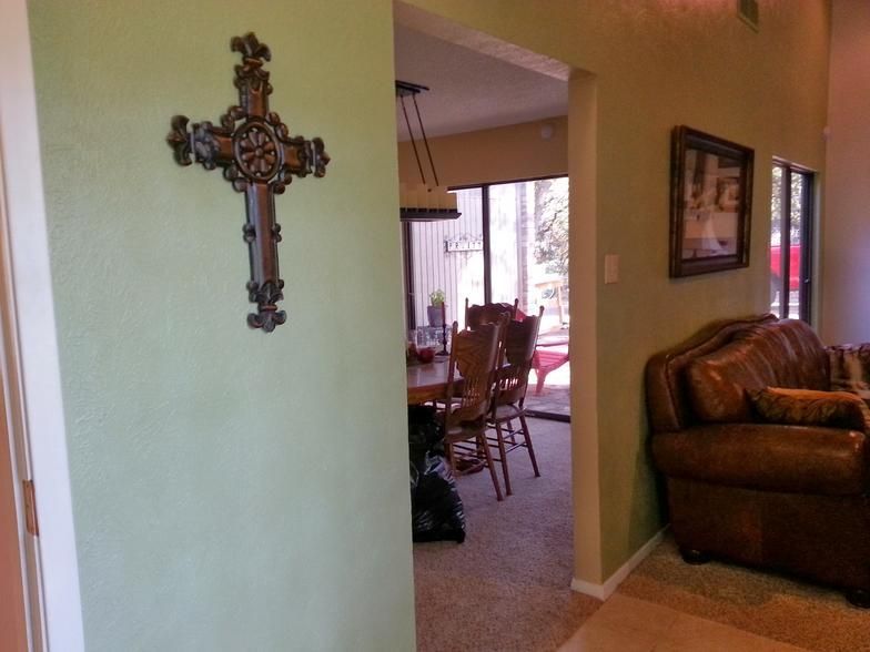 Ornate wooden cross on a green wall. Dining room with table and chairs visible through doorway. Leather sofa.