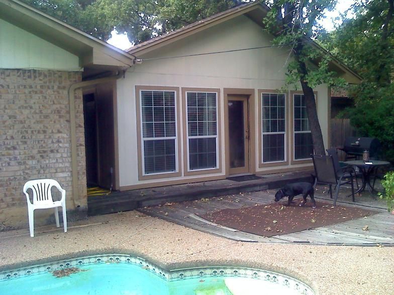Backyard patio with pool, building with windows and door. Dog on a mat, chair, table.