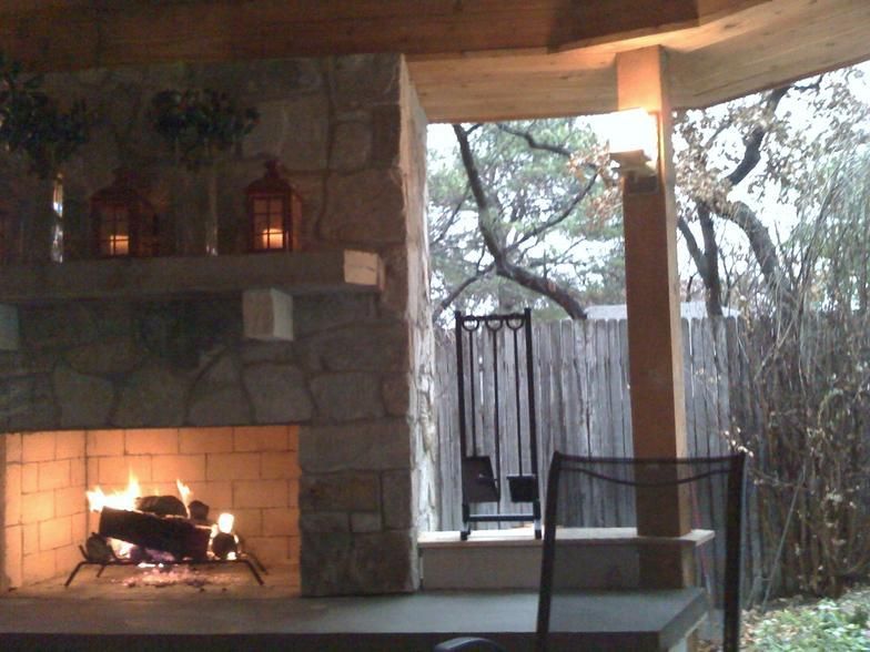Outdoor fireplace with burning logs, stone surround, lanterns, and view of a yard.