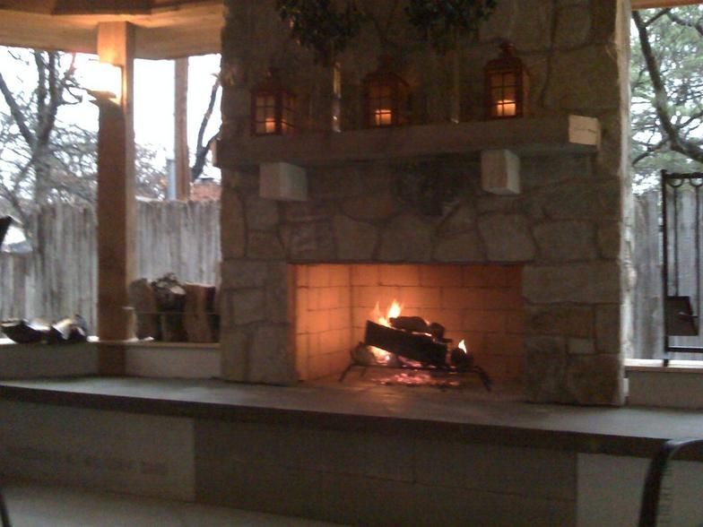 Stone fireplace with lit fire, lanterns, and wooden mantel on a patio.