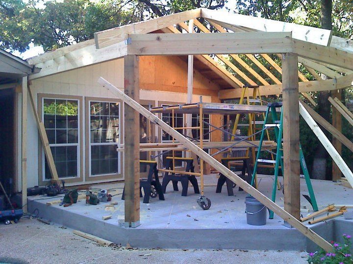 Construction of a wooden patio addition with exposed beams and scaffolding, next to a house with windows.