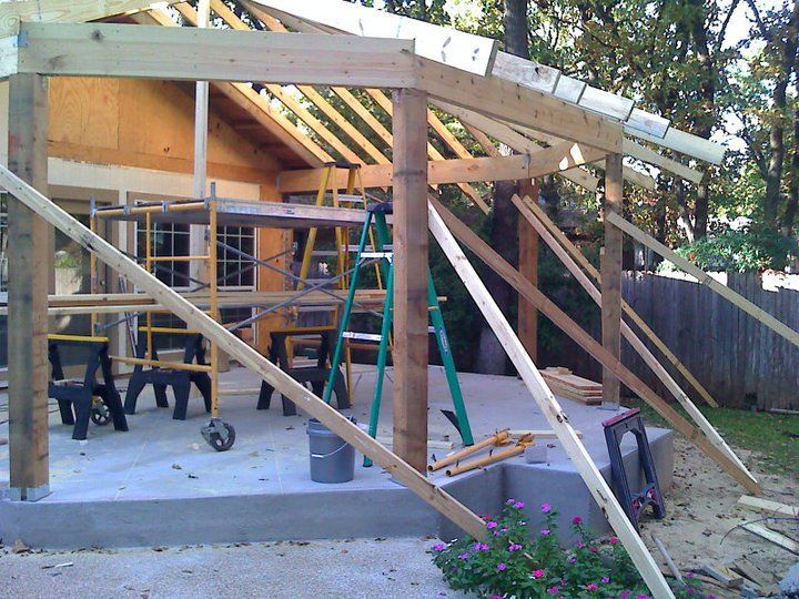 Construction of a wooden pergola attached to a house; supports and beams are in place on a concrete patio.