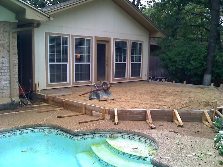Concrete patio construction next to a pool and a house with windows. Wooden forms define the patio's edges.