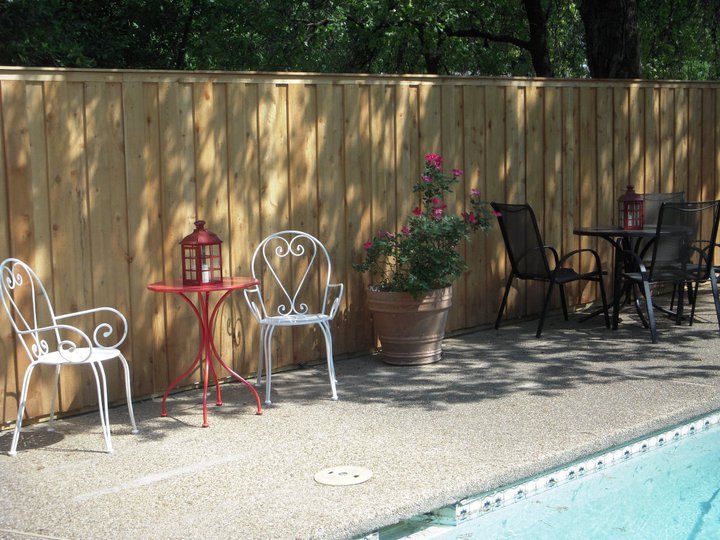 Wooden fence with patio furniture beside a pool. Red table, white chairs, potted roses.