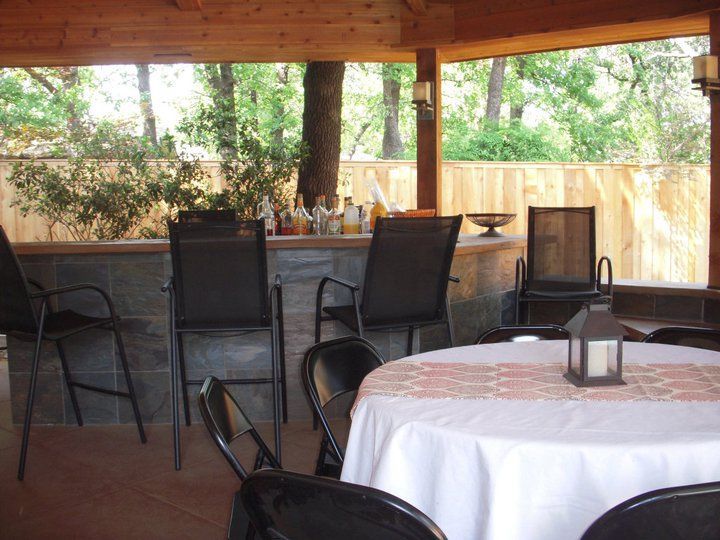 Outdoor bar with seating under a wooden structure, trees in the background, a round table with a tablecloth.