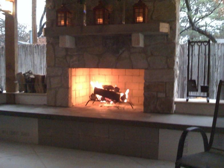 Stone fireplace with burning logs, outdoor setting. Lanterns on mantel, gray stone, wooden fence backdrop.
