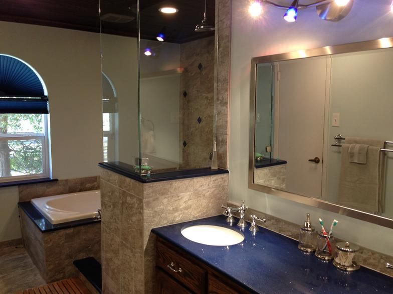 Bathroom with blue countertop, marble-like wall, tub, glass shower, arched window with blue shade, and mirror.