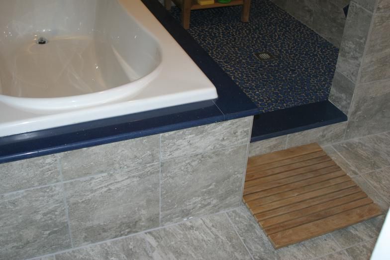Bathroom with a white bathtub, blue trim, and gray tile. A wooden bath mat sits on the floor.
