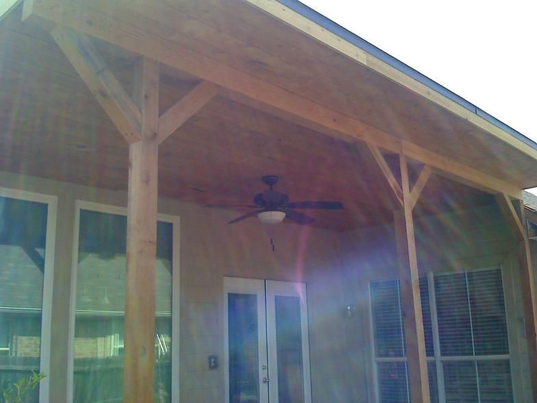 Covered patio with wooden supports, ceiling fan, and access to a house with windows and doors.