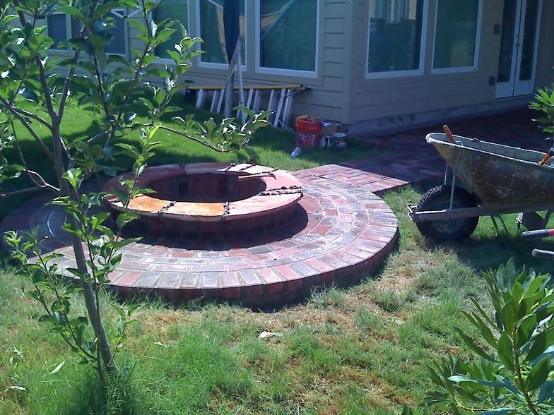 Brick fire pit in a grassy backyard with a path leading to it.