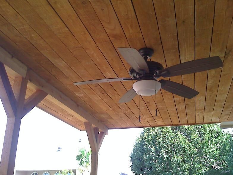 Wooden patio ceiling with a dark ceiling fan and a view of trees.