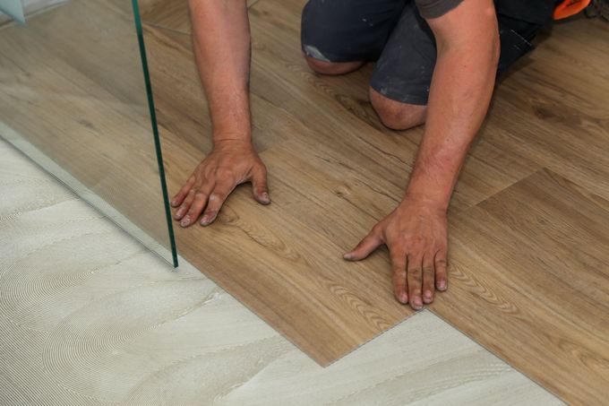 Person installing wood-look flooring next to a glass shower panel.