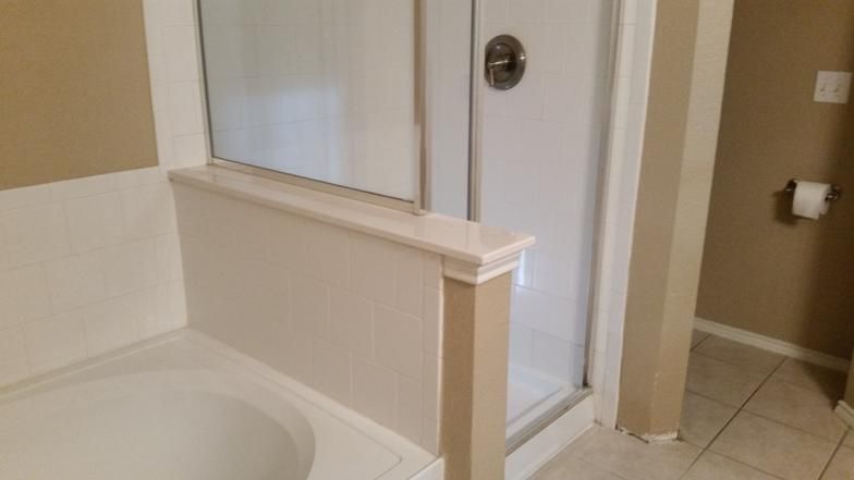 Bathroom with a white bathtub and shower. Beige walls, sliding glass door, and a roll of toilet paper are visible.