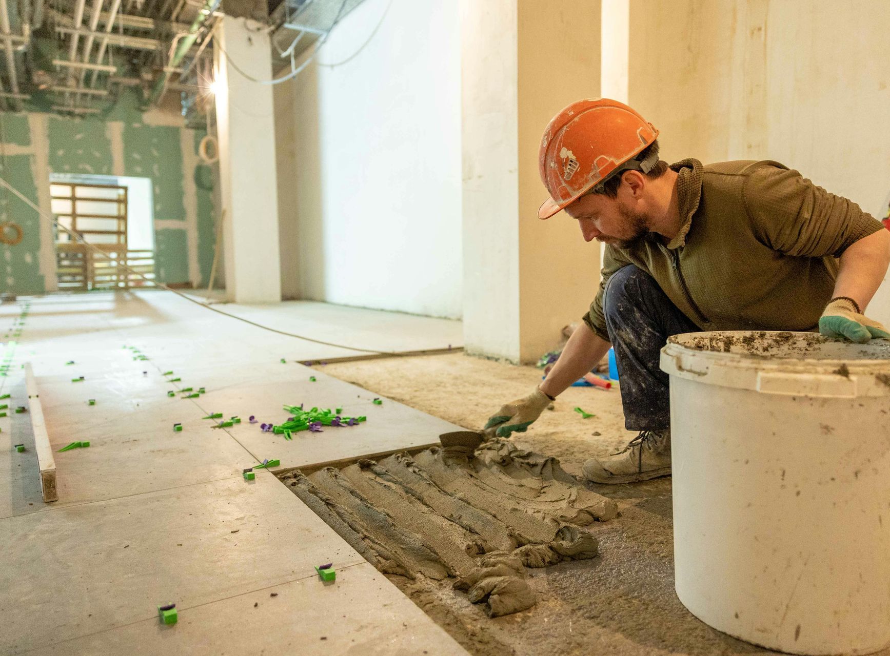 A man in a hard hat is laying tiles on a floor.