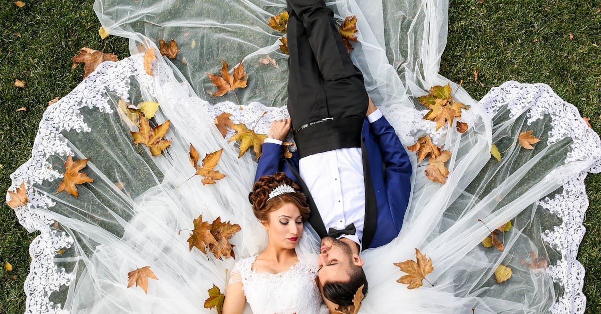 A bride and groom are laying on their backs in the grass covered in leaves.