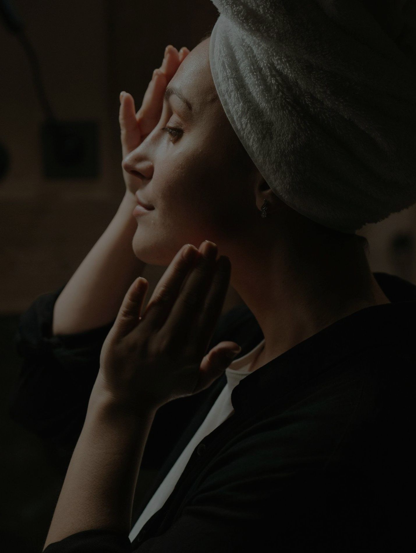 A woman with a towel wrapped around her head is looking at her face.