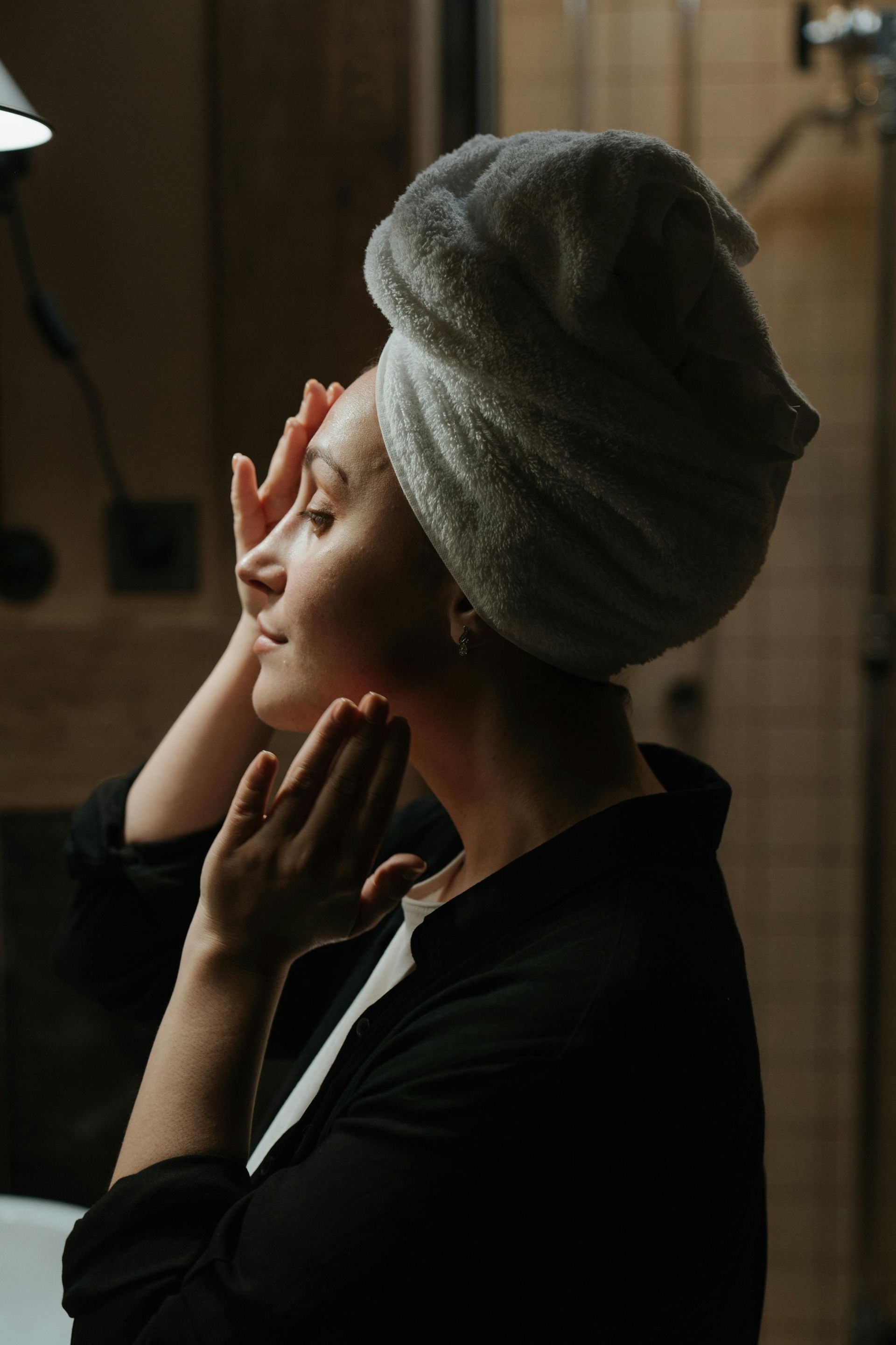 A woman with a towel wrapped around her head is looking at her face in the mirror.