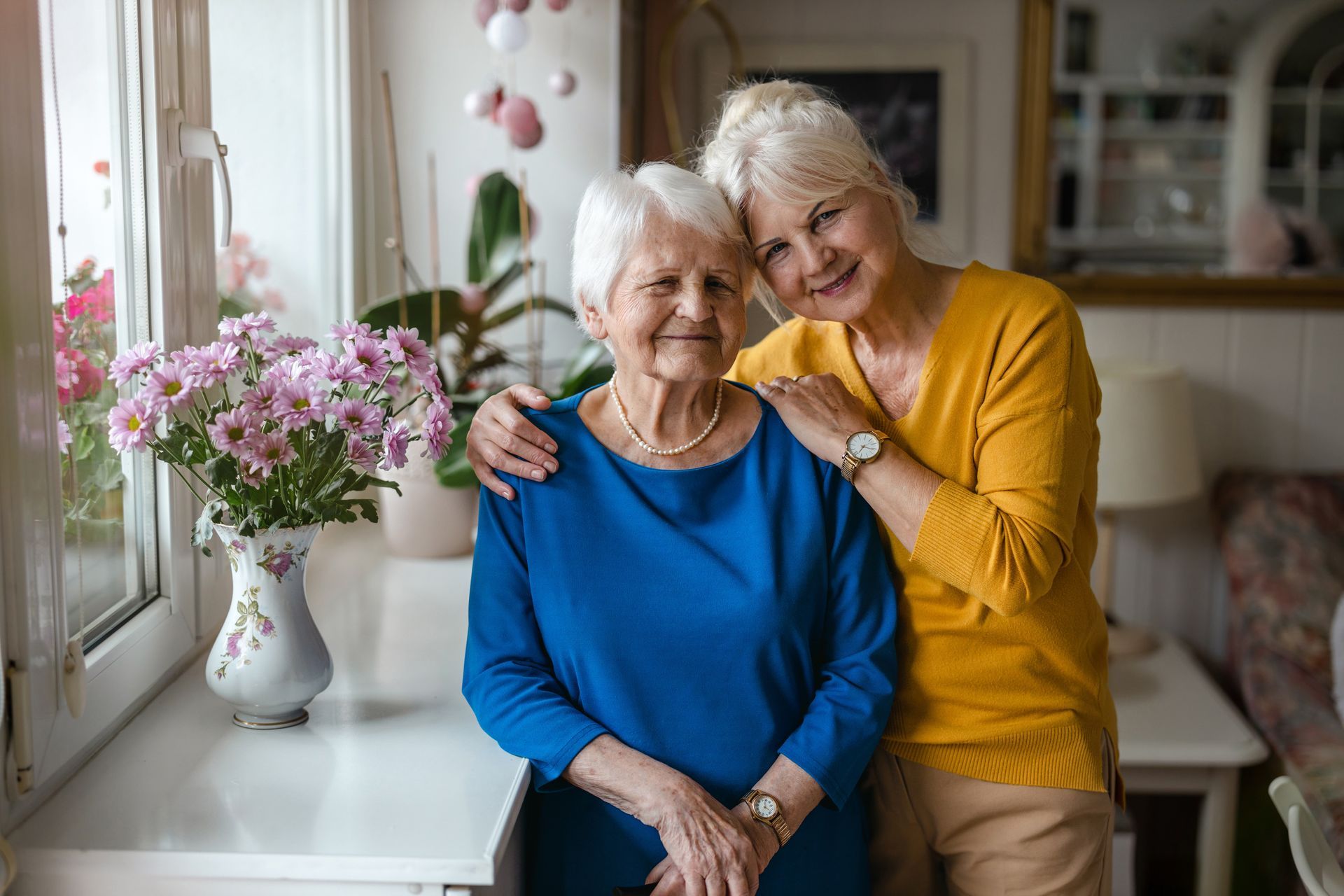 Two older women are posing for a picture in front of a window.