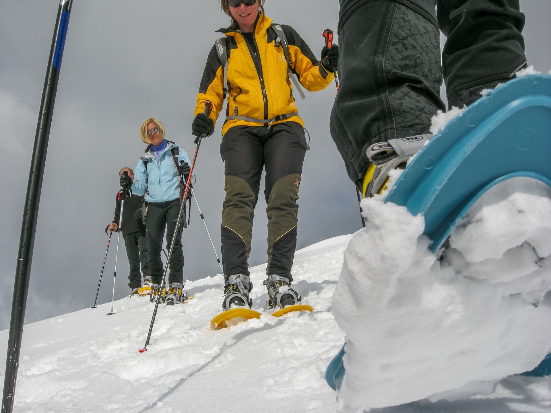 Eine Frau in einer gelben Jacke läuft mit Schneeschuhen auf einem schneebedeckten Hügel.