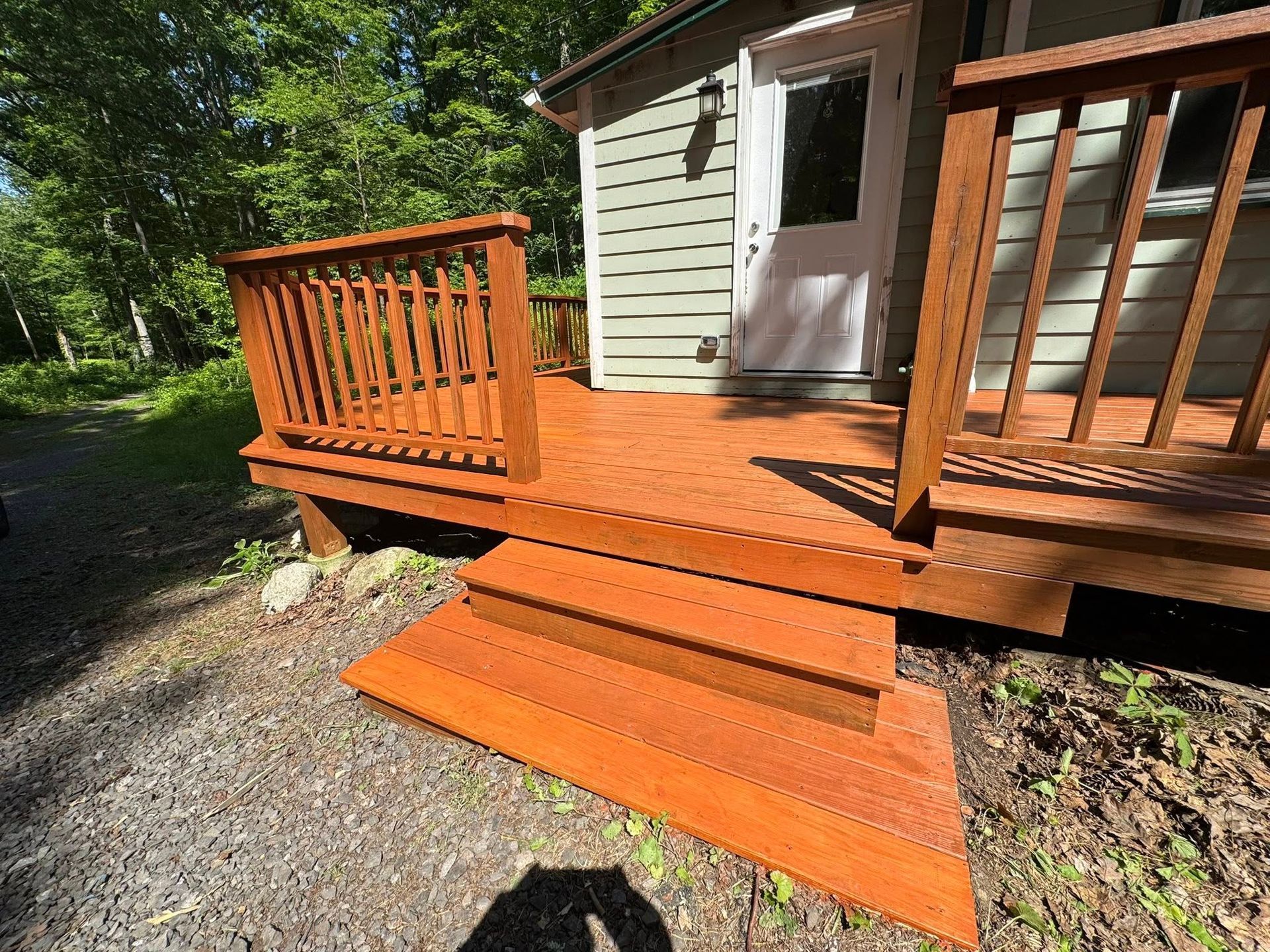 Wooden deck and steps painted reddish-brown leading to a small cabin with light green siding and a white door; sunny outdoor setting.