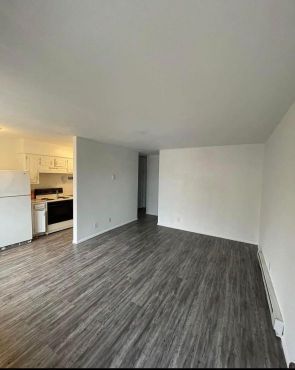 An empty living room with hardwood floors and a kitchen in the background.