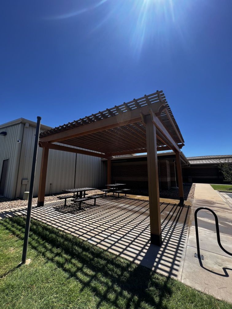 A wooden pergola with picnic tables underneath it in front of a building.