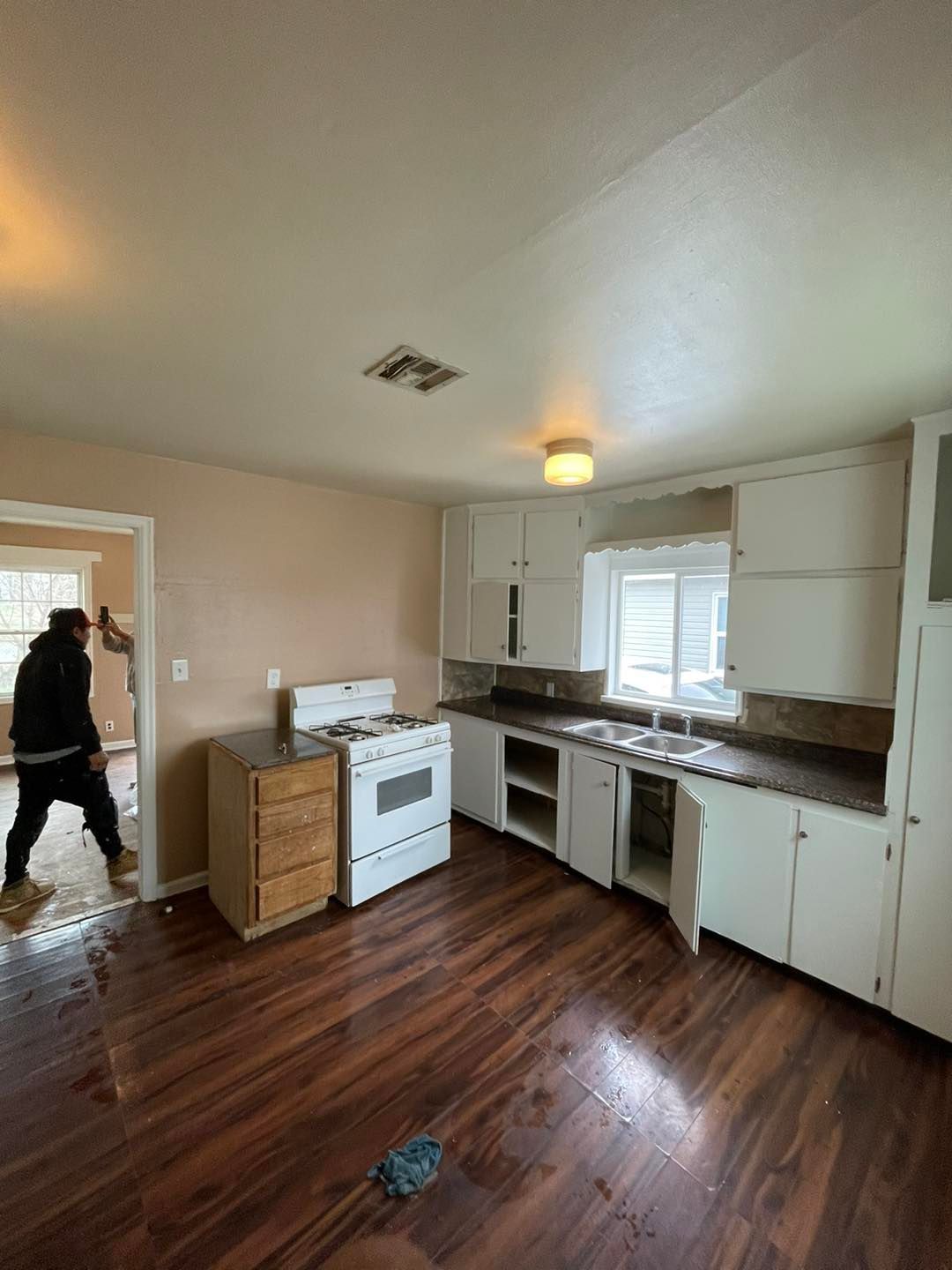 A man is standing in an empty kitchen with wooden floors and white cabinets.