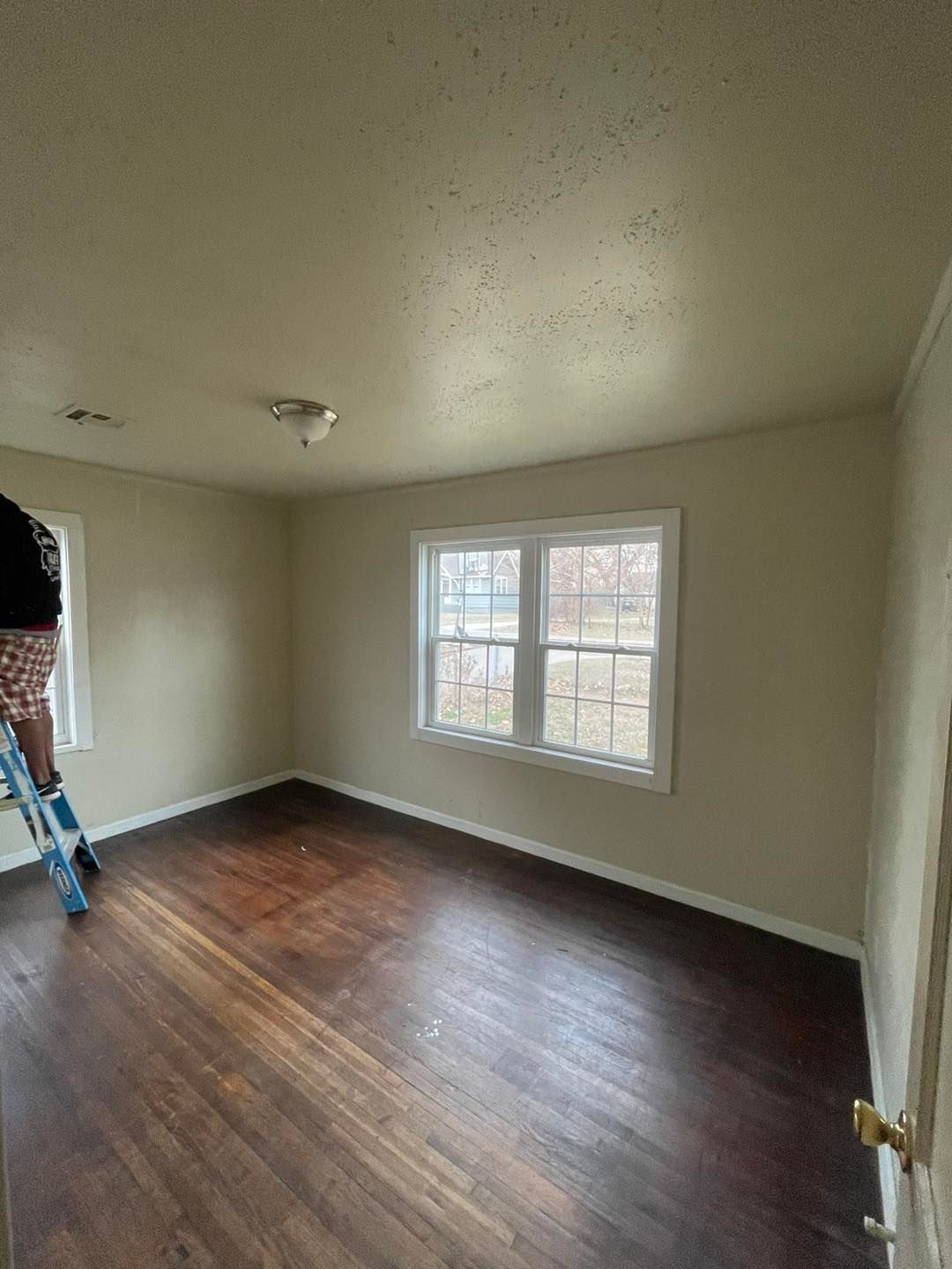 A person is standing on a ladder in an empty room with hardwood floors.