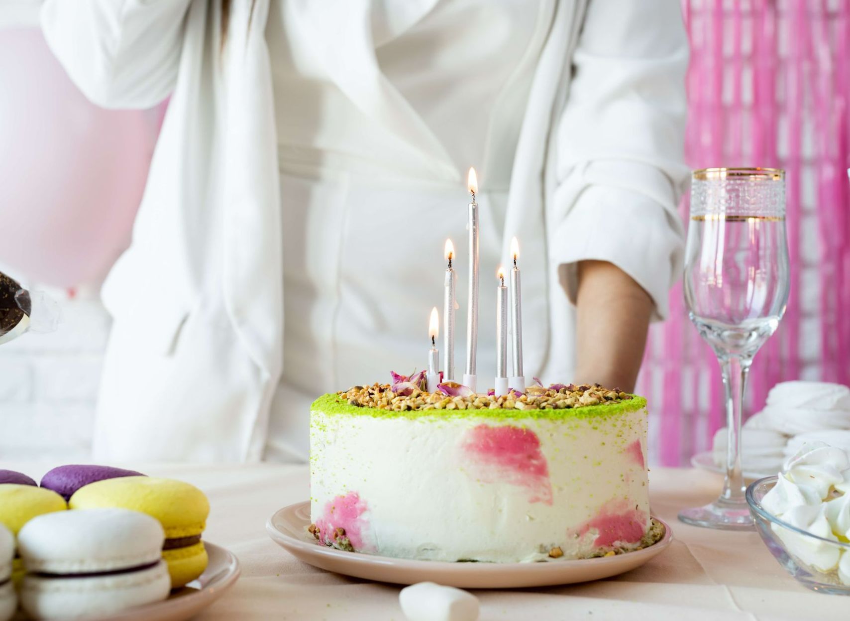 Birthday cake with lit candles, surrounded by macarons, a drink and person in white blazer.