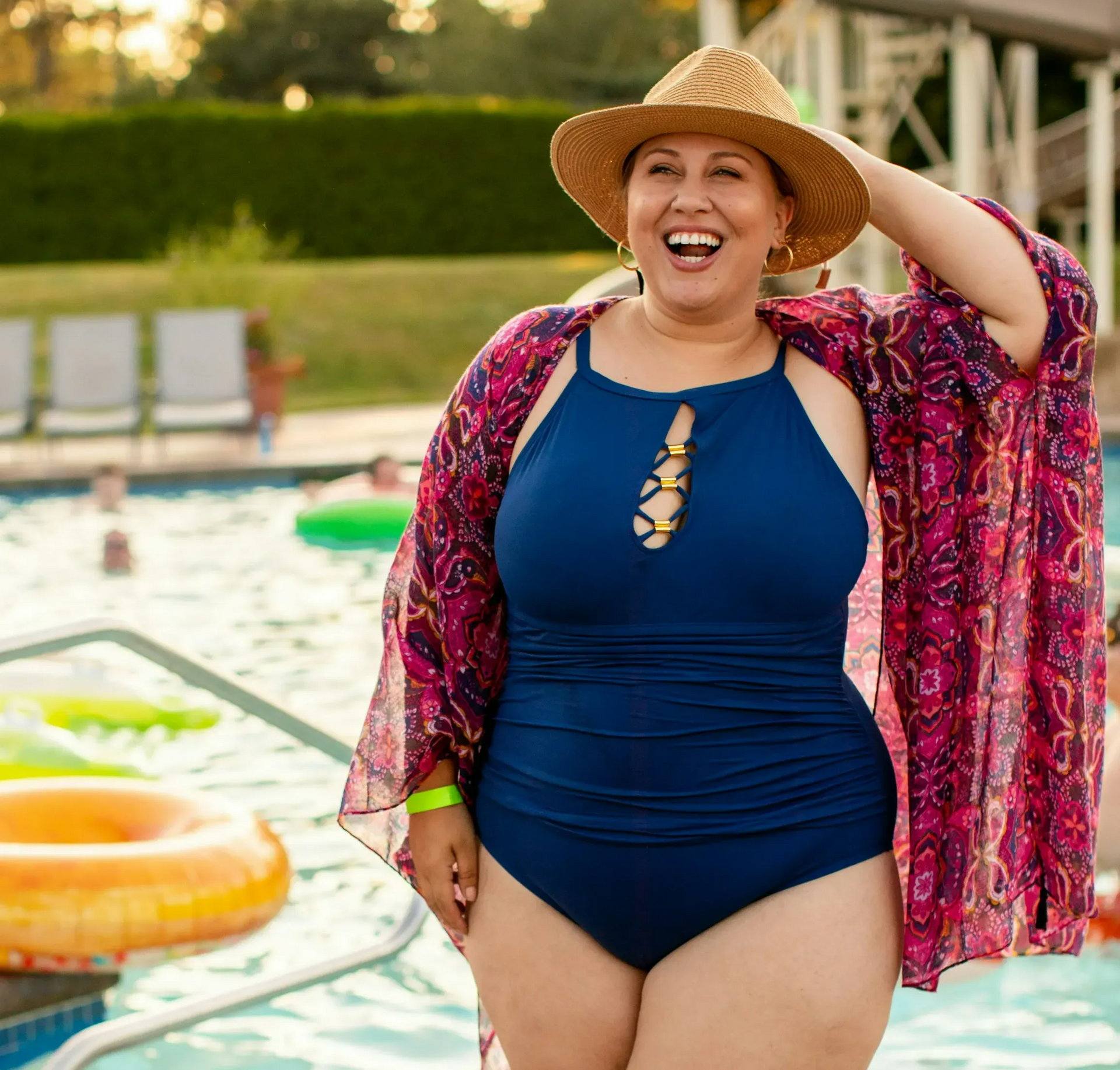 Woman in a navy swimsuit smiles by a pool, wearing a straw hat and colorful cover-up.