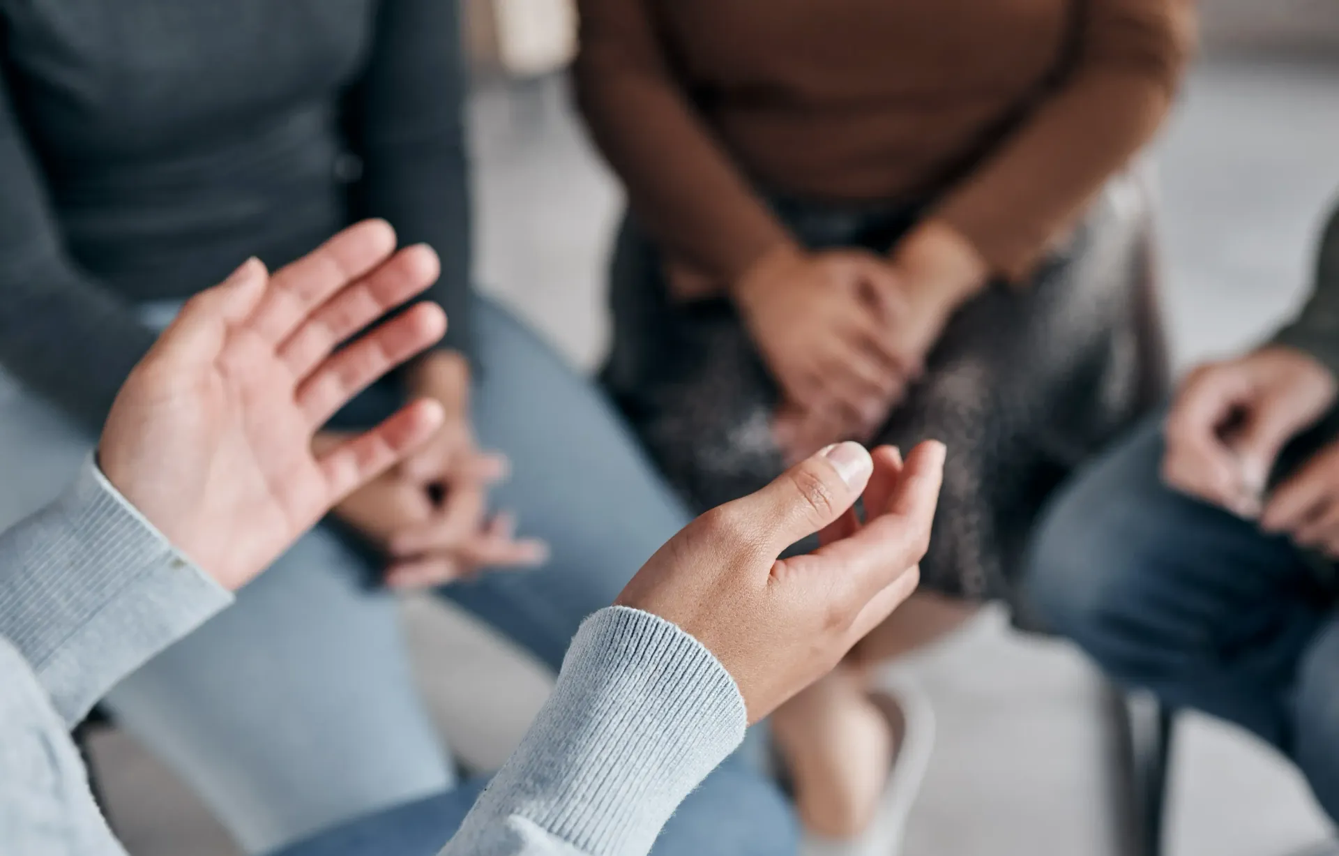 Hands gesturing, speaking in a group therapy setting. Other people are seated nearby, listening.