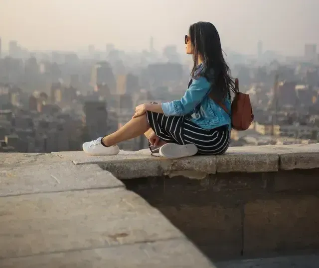 Woman seated on ledge, looking at a cityscape, wearing sunglasses, blue jacket, striped skirt, backpack.