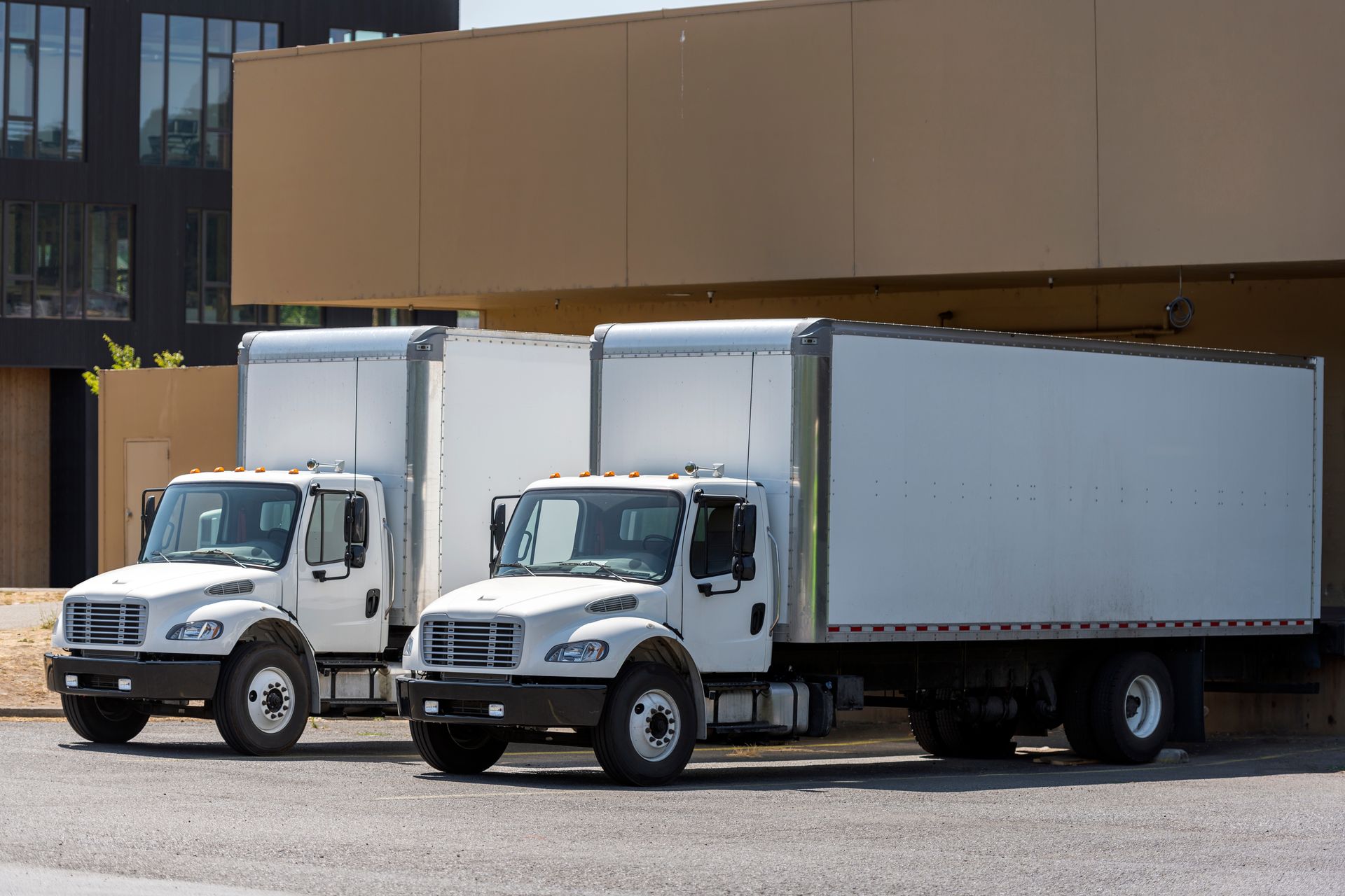 Two white box trucks parked in front of a tan building.