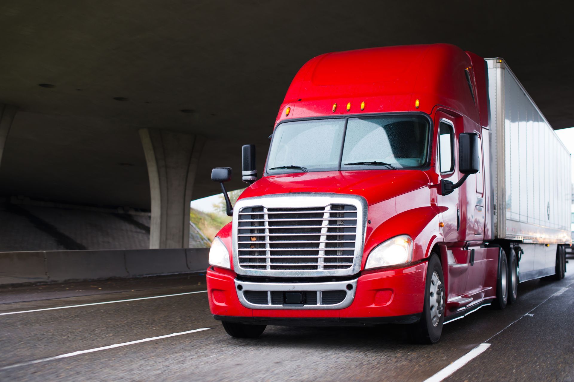 Red semi-truck driving on a highway, under a concrete overpass.