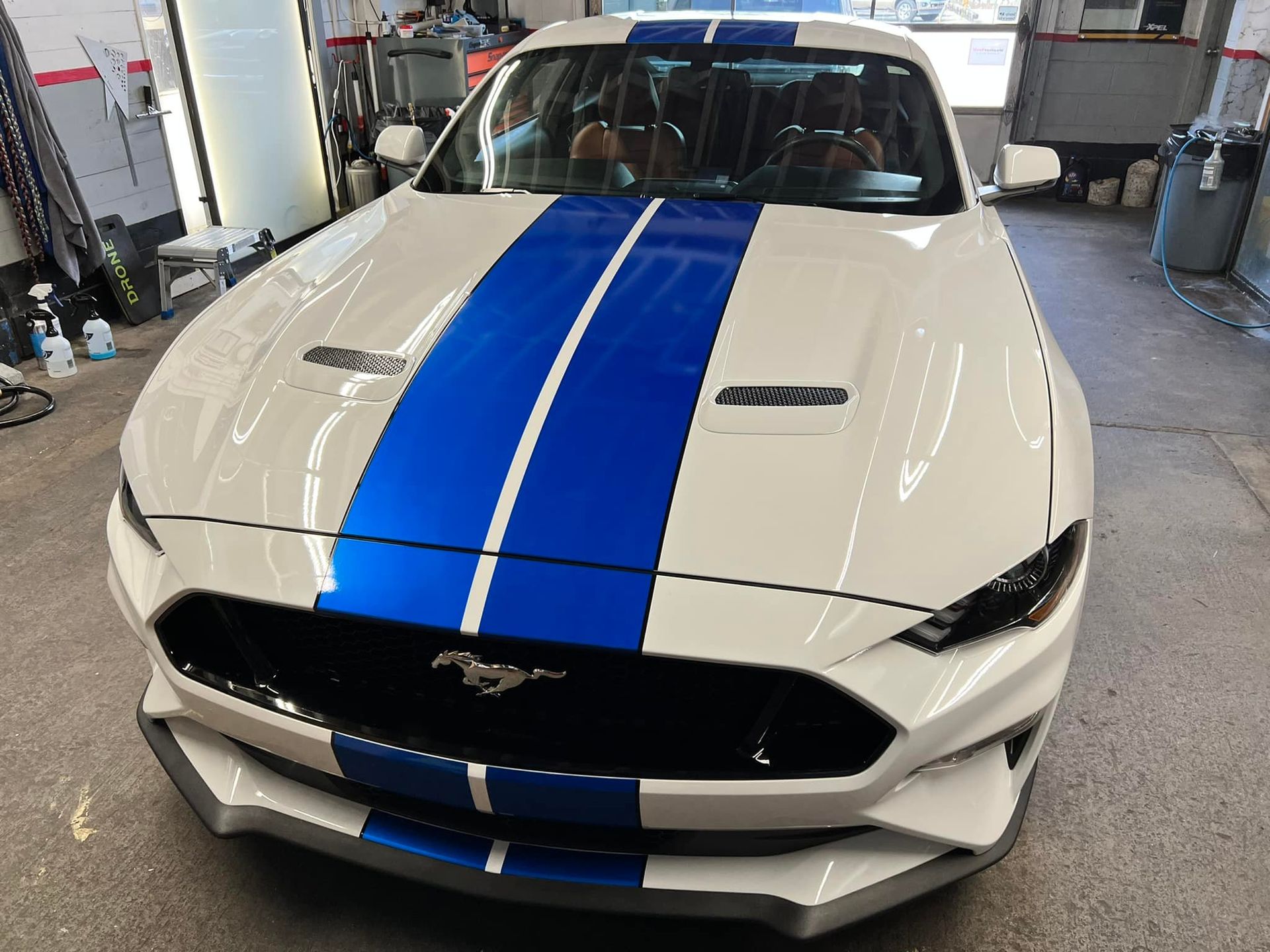 A white mustang with blue stripes on the hood is parked in a garage.