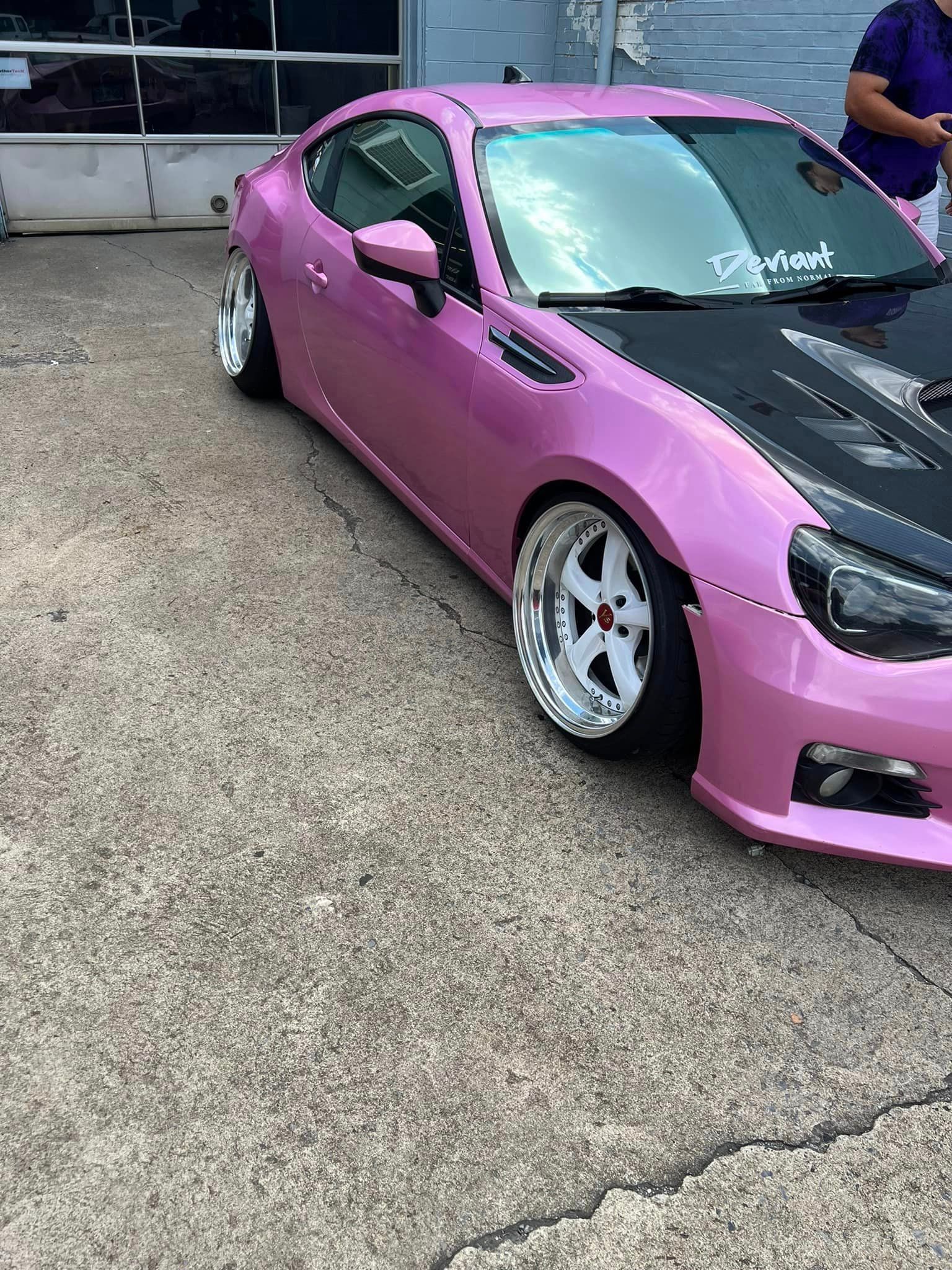 A pink car with a black hood is parked in a parking lot.