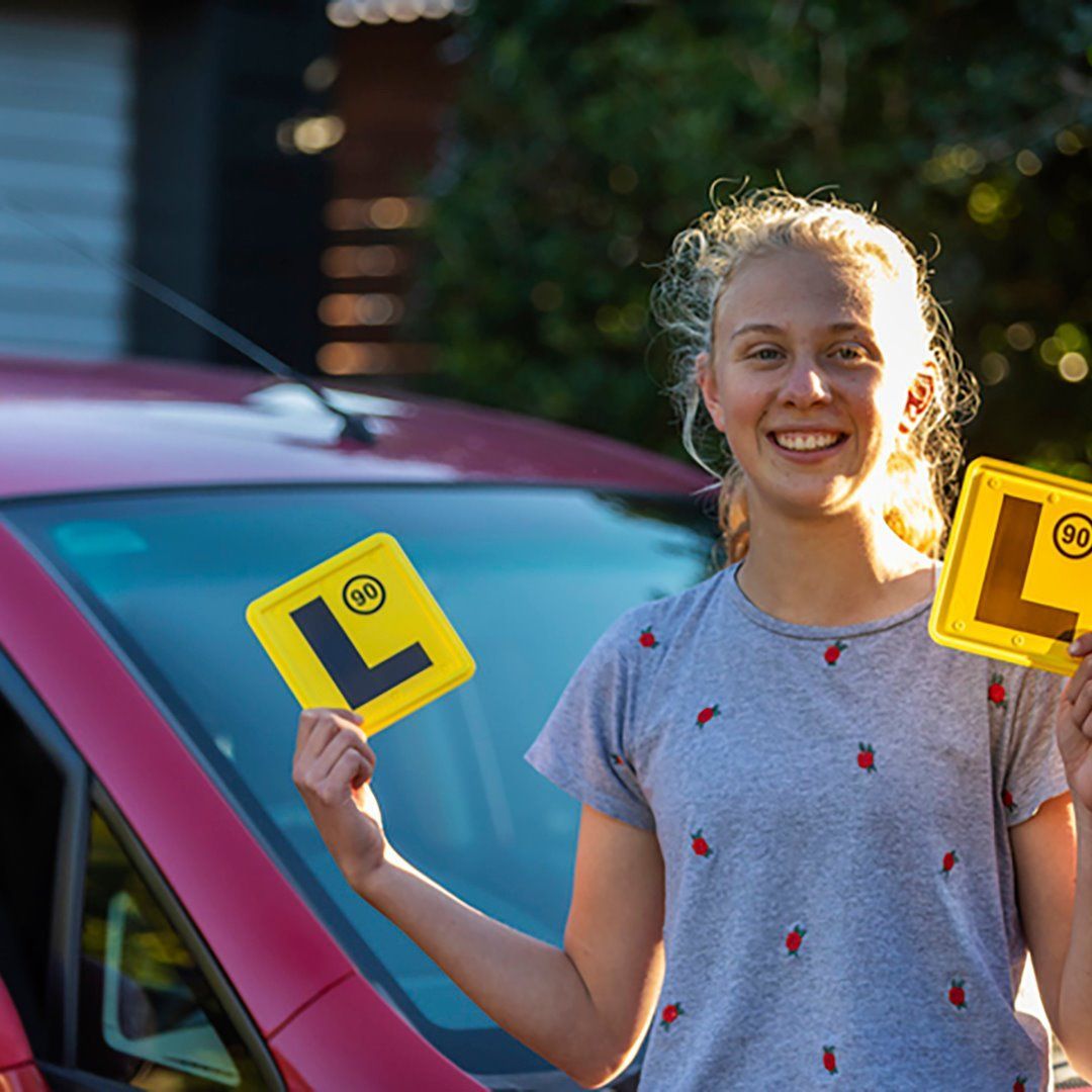 A Woman is Holding Two Yellow L Signs in Front of a Pink Car — Ian Emslie Optometrist In Warners Bay, NSW