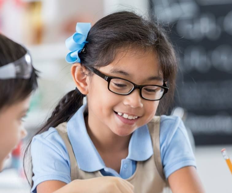 A Young Girl Wearing Glasses is Sitting at a Desk in a Classroom — Ian Emslie Optometrist In Warners Bay, NSW