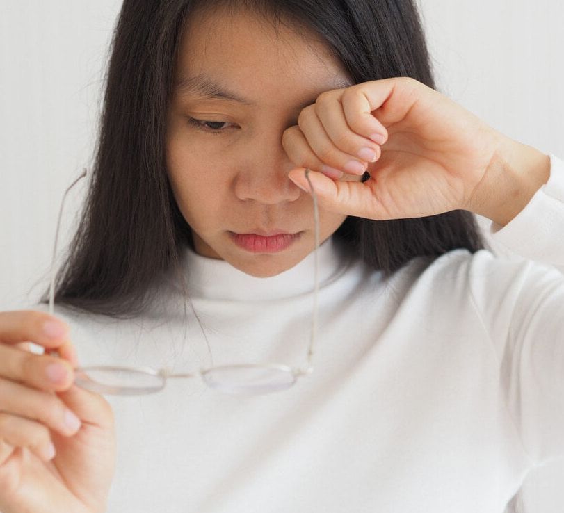 A Woman is Rubbing Her Eyes While Holding a Pair of Glasses — Ian Emslie Optometrist In Warners Bay, NSW
