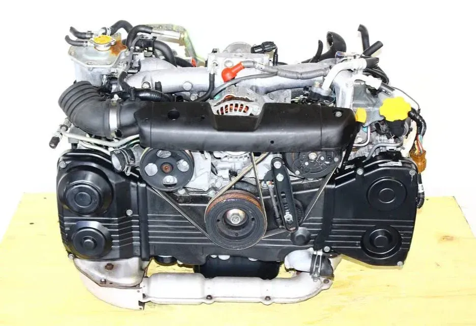 A Subaru flat-four engine shown from the front, featuring black timing covers, pulleys, and metallic intake components.