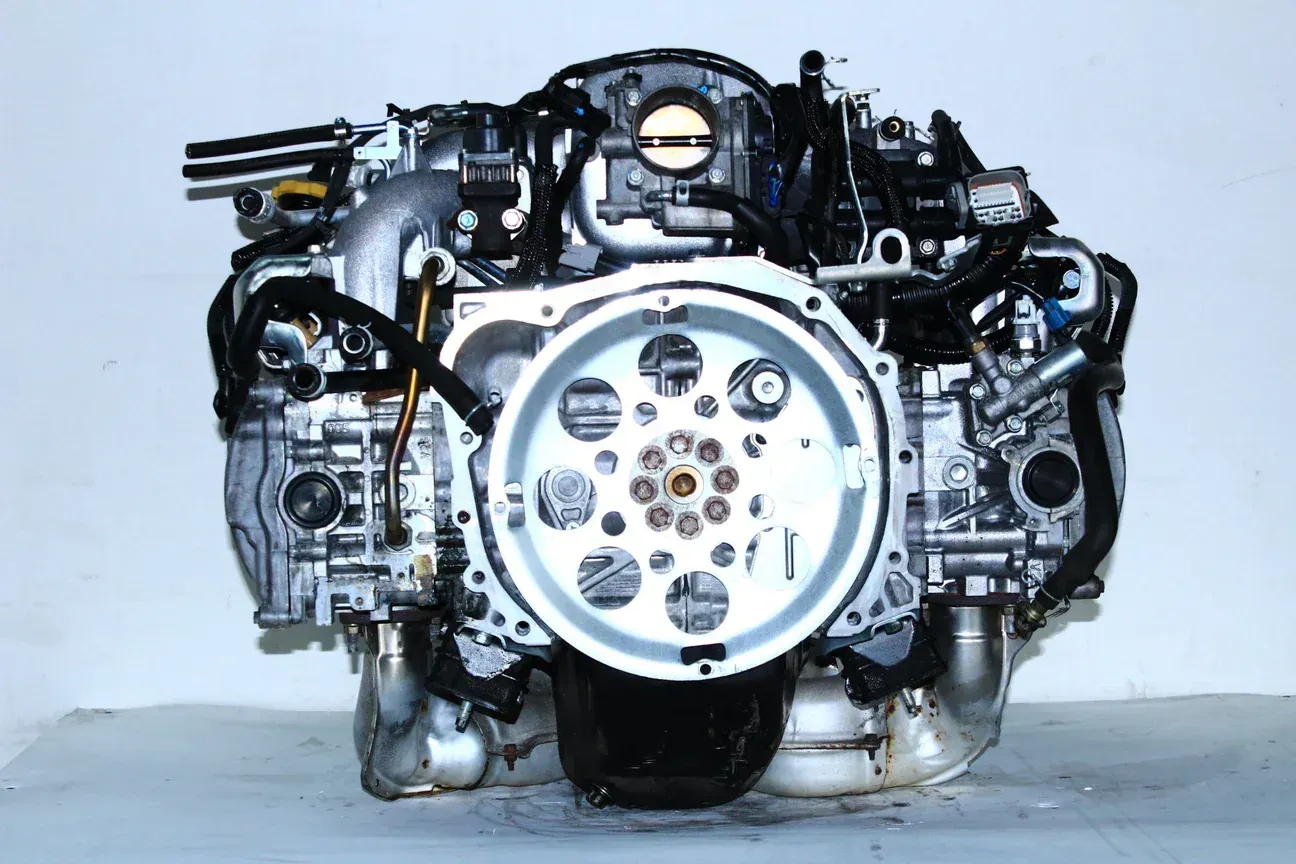 A silver automotive boxer engine viewed from the rear, showcasing its flywheel and various intake and electrical components.
