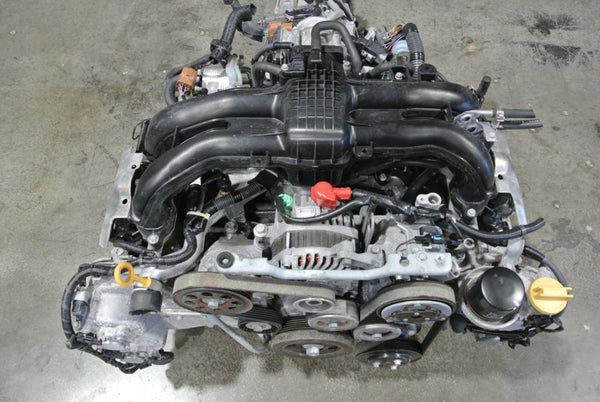 A clean, overhead view of a Subaru boxer engine sitting on a gray concrete floor, showing intake, pulleys, and belt.