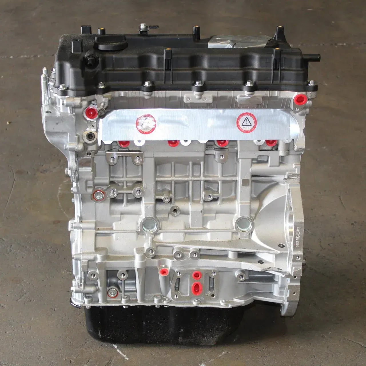 A silver automobile engine block with a black top cover, placed on a dark floor with visible red port caps.