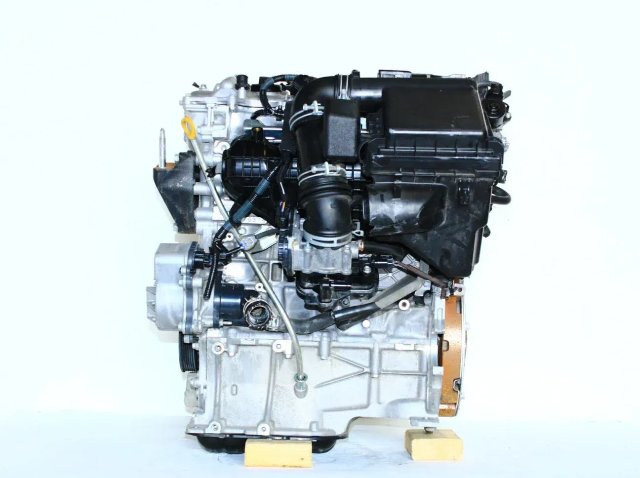 A detached, silver automotive engine with a black intake manifold, resting on two small wooden blocks against a white wall.