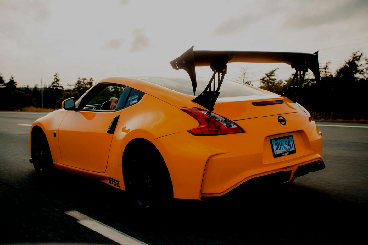 Bright orange Nissan 370Z sports car with a large rear wing driving on a road during the daytime.