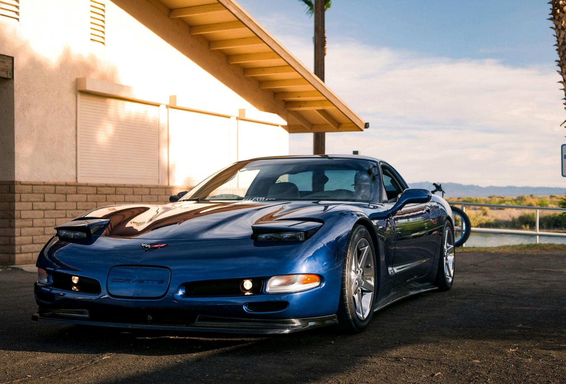 A dark blue C5 Chevrolet Corvette parked on an asphalt lot beside a building during a sunny day.