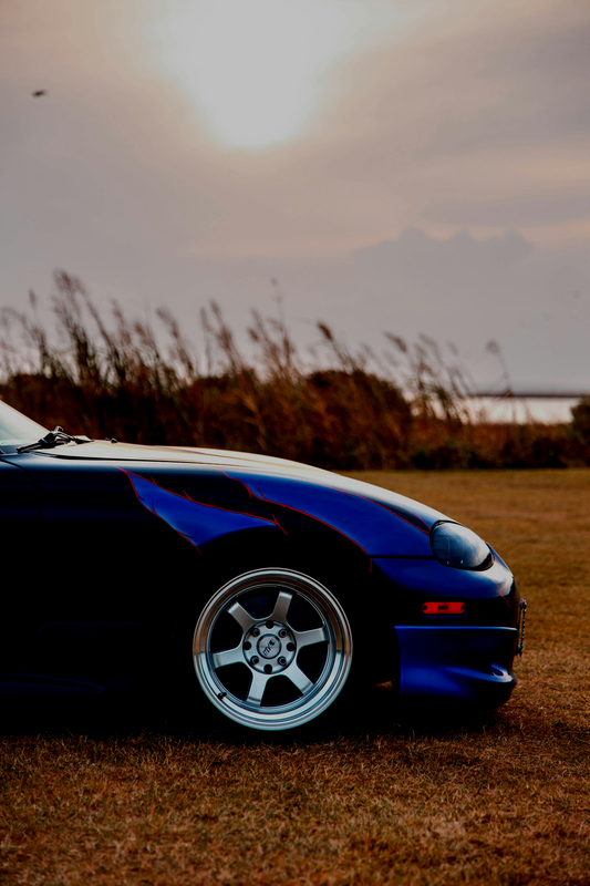 A blue sports car parked on a grassy field at sunset, with tall reeds in the background.